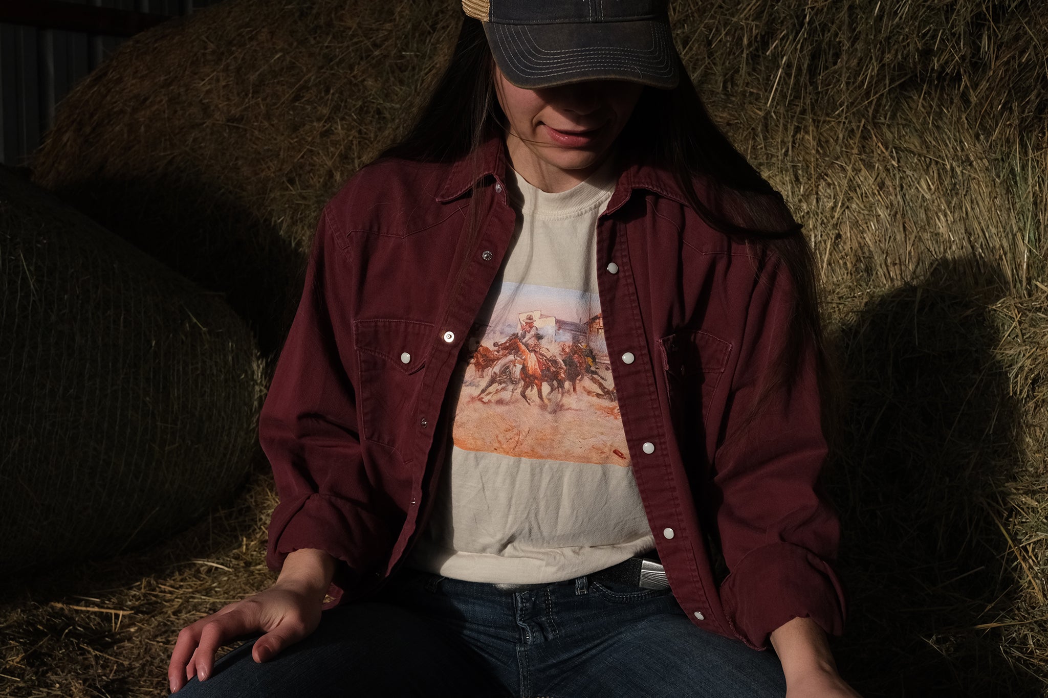 Person wearing a maroon flannel over a graphic C.M. Russell t-shirt sitting on hay.