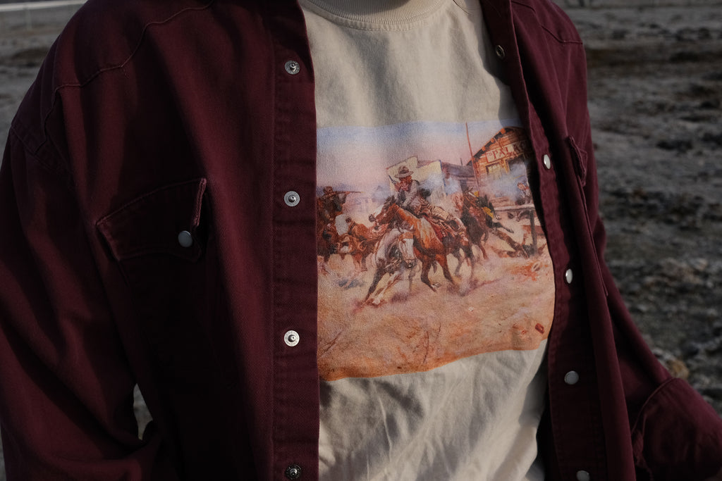 Beige T‑shirt featuring a cowgirl wearing a Charles Marion Russell’s Smoke of a .45 t-shirt depicting a cowboy firing his revolver in a tense Western scene.