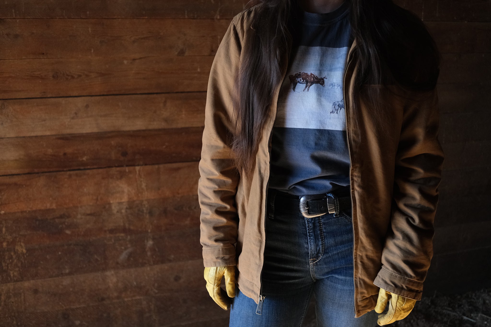 Women wearing a brown jacket, blue jeans, and yellow gloves against a wooden wall.