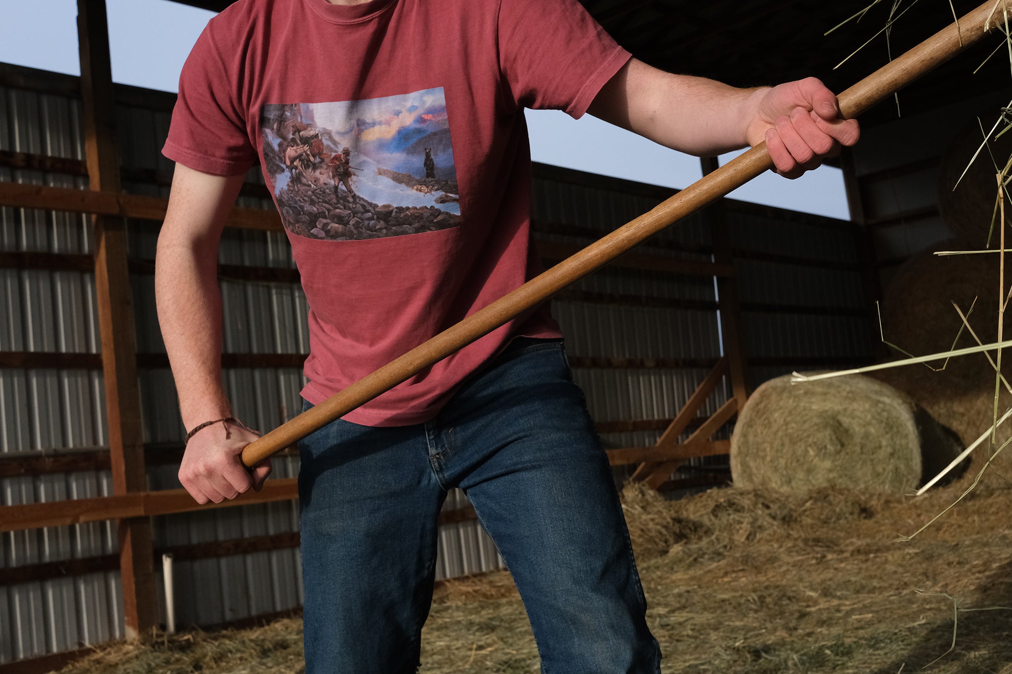 Person in a barn scooping hay, wearing a red t-shirt with a C.M. Russell graphic T-shirt design.